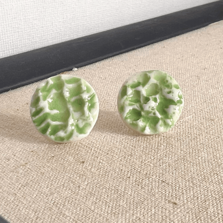 Porcelain sea pebble stud earrings on white canvas background with green marbled glaze pattern.