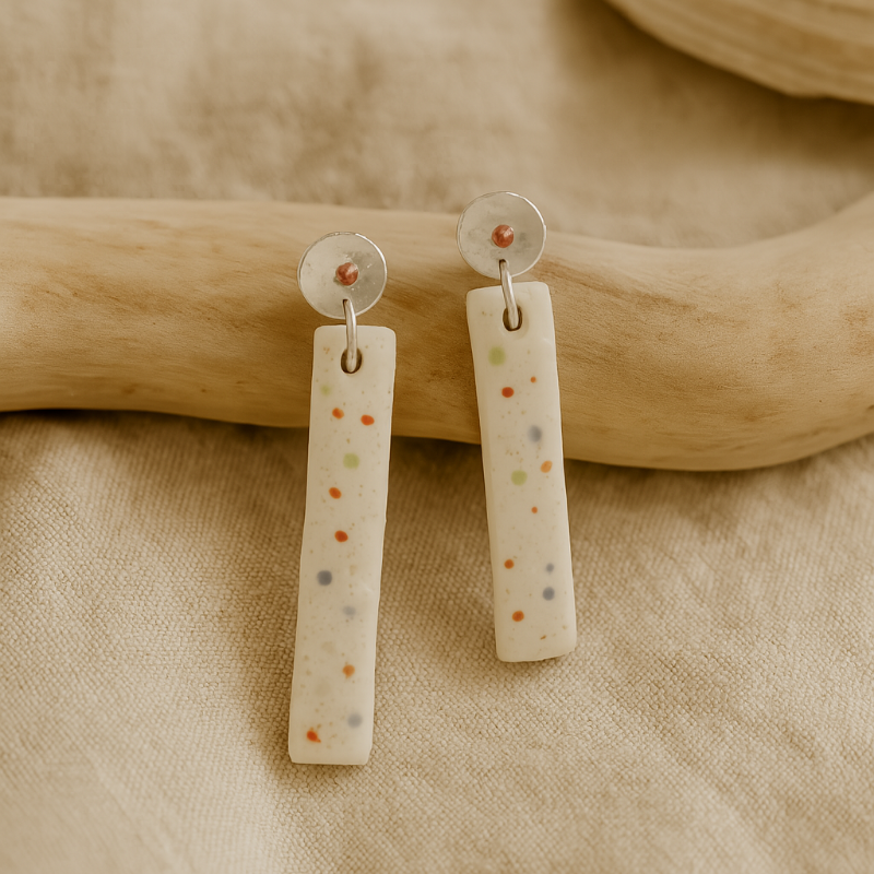Pair of handmade porcelain earrings with subtle multicolour dots and sterling silver and copper beaten stud tops, photographed against a linen background.