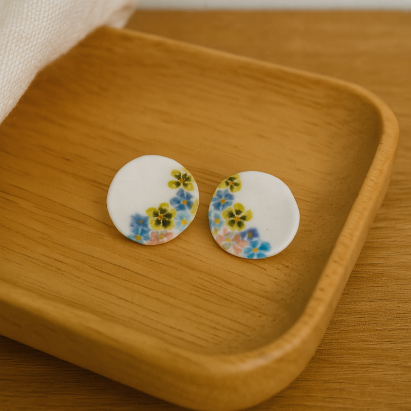 Round hand-painted porcelain earrings with pink, blue, and yellow floral designs on a wooden background.