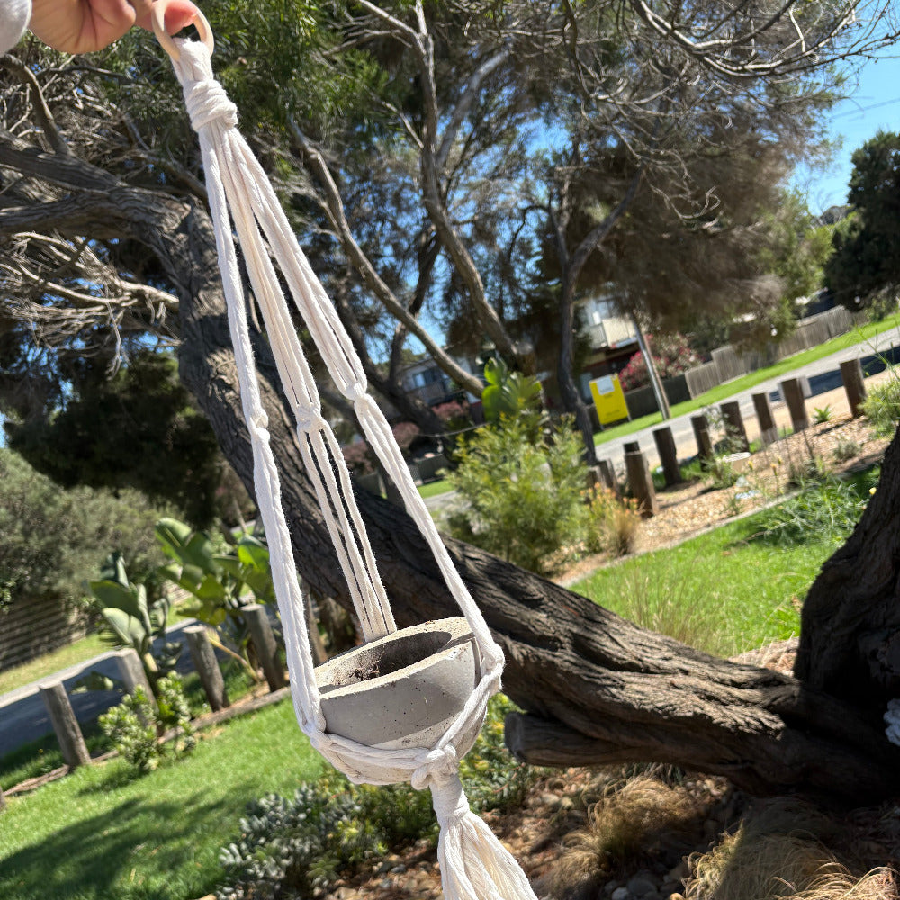 White macrame hanger hanging from a tree in an outdoor setting with grass and trees in the background.