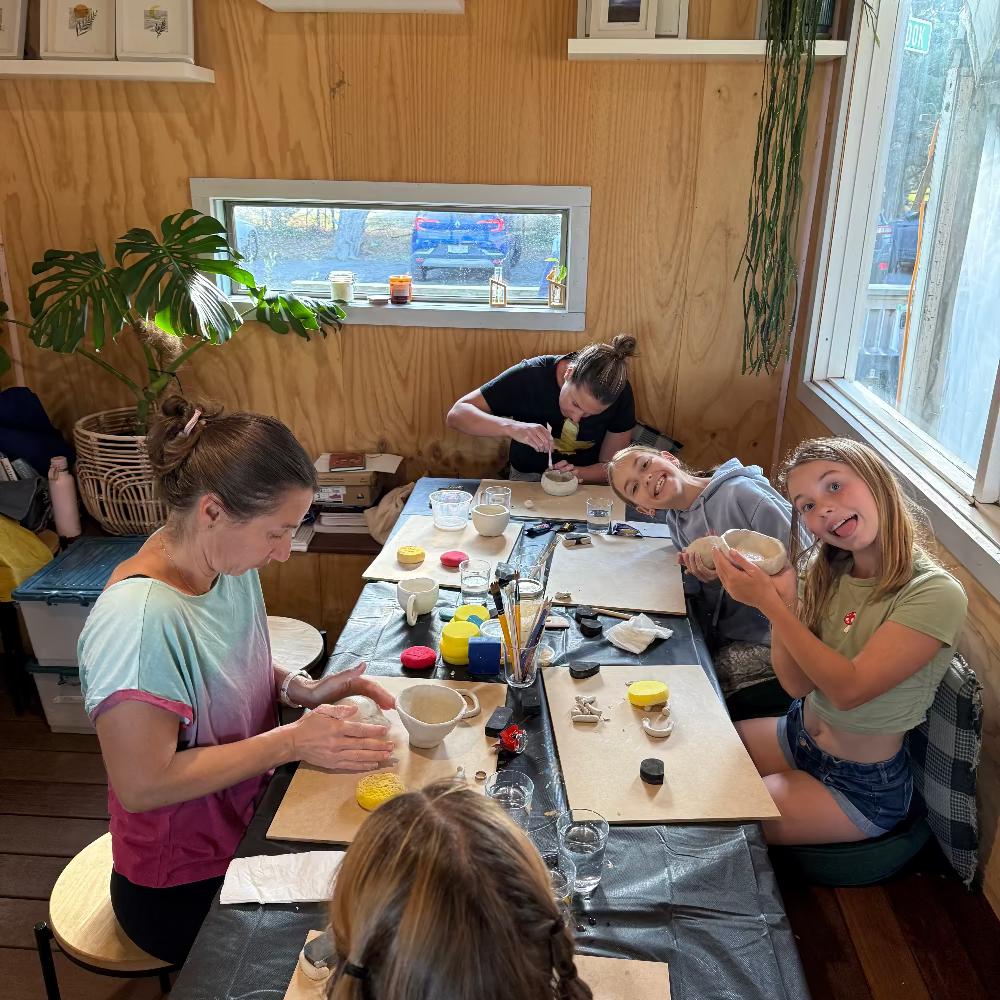 Group of people sitting around a table in a cozy room with wooden walls and large windows doing a clay workshop.