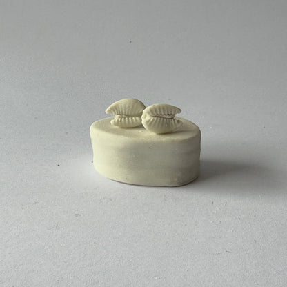 Handmade porcelain cowrie shell stud earrings in natural white, photographed on a ceramic pedestal.