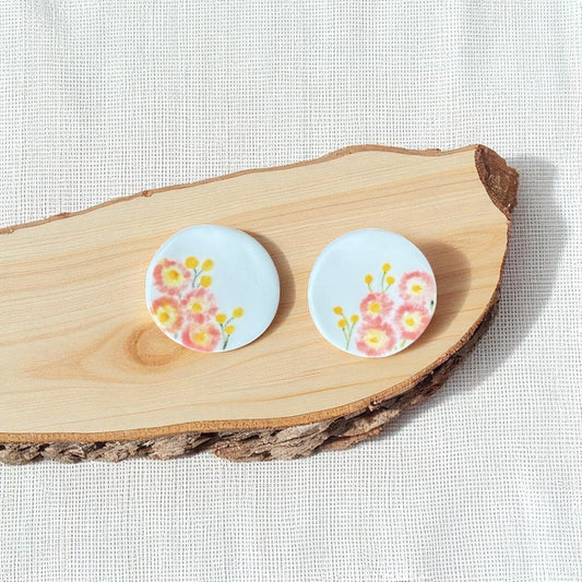 Porcelain stud earrings hand-painted with pink paper daisies and yellow billy buttons, displayed on natural wood.