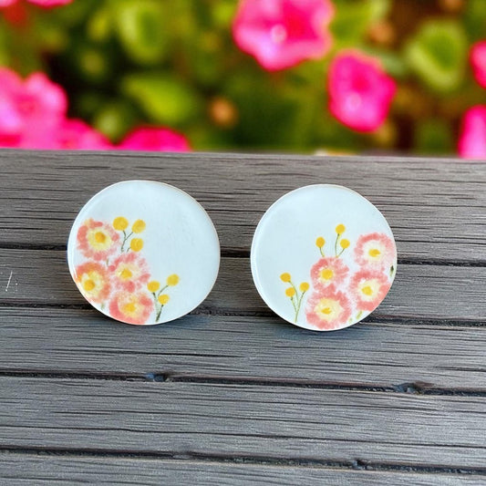 Porcelain stud earrings hand-painted with pink paper daisies and yellow billy buttons, displayed on natural wood.