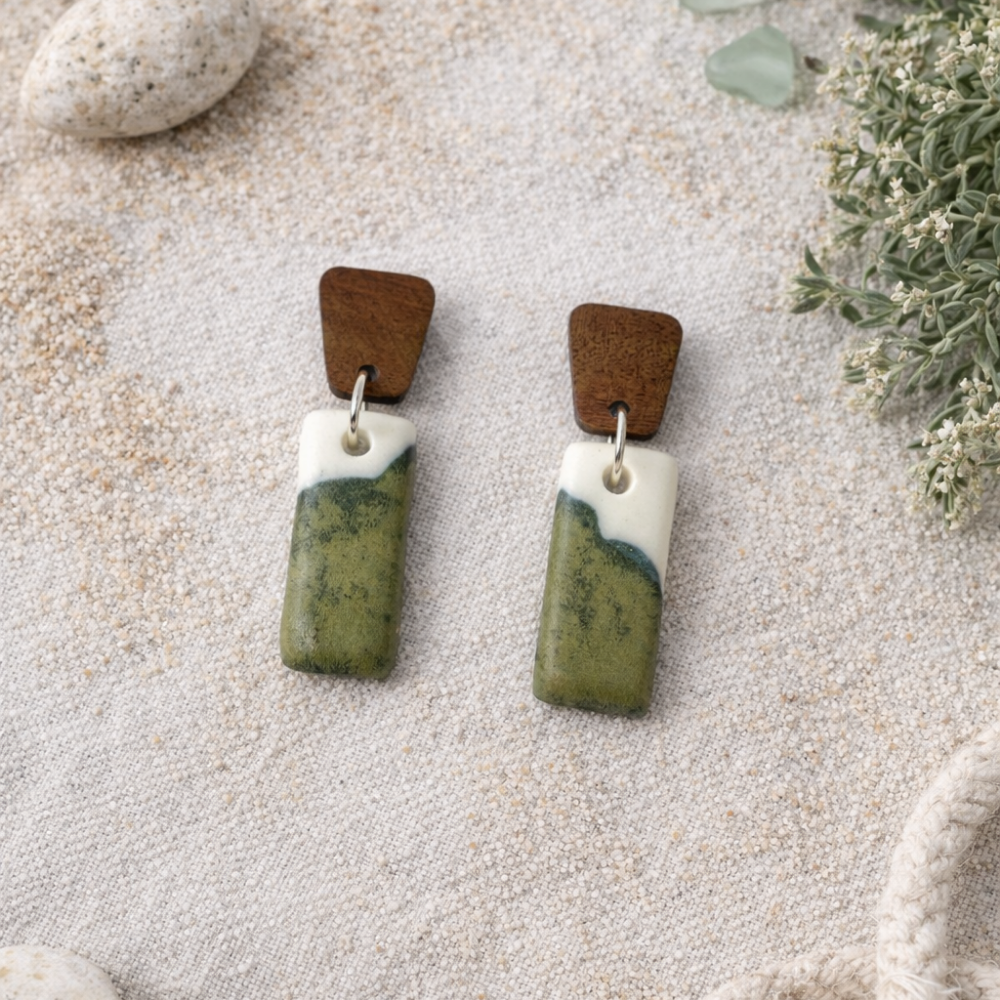 Handmade Inlet porcelain dangle earrings with green and ivory glaze and wooden studs on a coastal bush flatlay background.