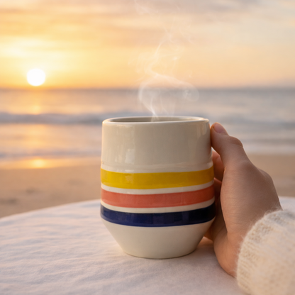 Hand holding a striped mug with a sunsrise over water in the background