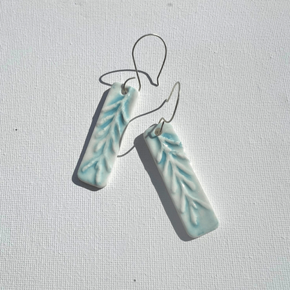 A pair of rectangular porcelain earrings in baby blue with sea leaf textures, resting on white canvas with handmade sterling silver hooks.
