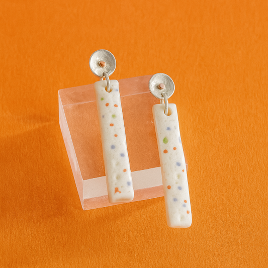 Pair of handmade porcelain earrings with subtle multicolour dots and sterling silver and copper beaten stud tops, photographed against an orange background.

