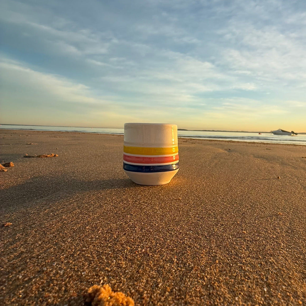 Handmade porcelain cup with bright sunrise stripes sitting on sand at sunrise, symbolising joy and a new day.