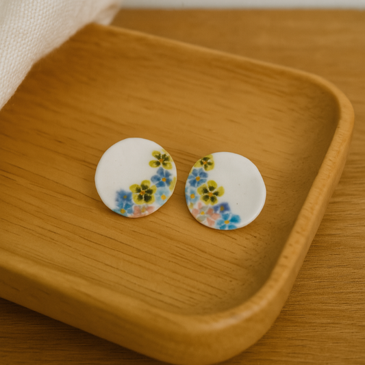 Round hand-painted porcelain earrings with pink, blue, and yellow floral designs on a wooden background.