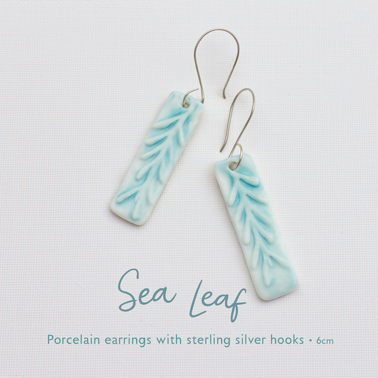 A pair of rectangular porcelain earrings in baby blue with sea leaf textures, resting on white canvas with handmade sterling silver hooks.