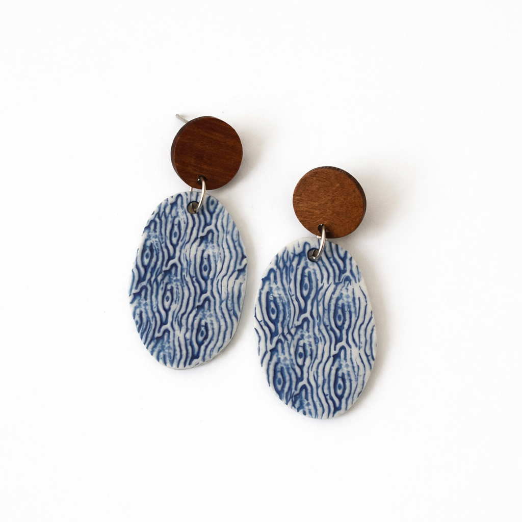 Handmade oval porcelain dangle earrings with blue and white sea-inspired pattern and wooden studs on a white background
