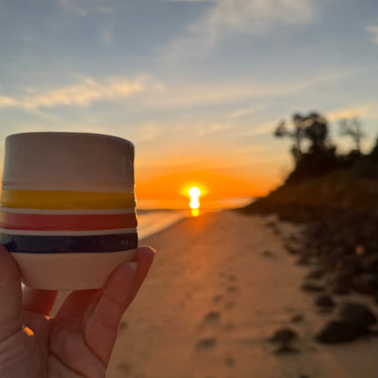 Handmade porcelain sunrise cup with colourful stripes, coastal ceramic mug photographed on beach at sunrise.